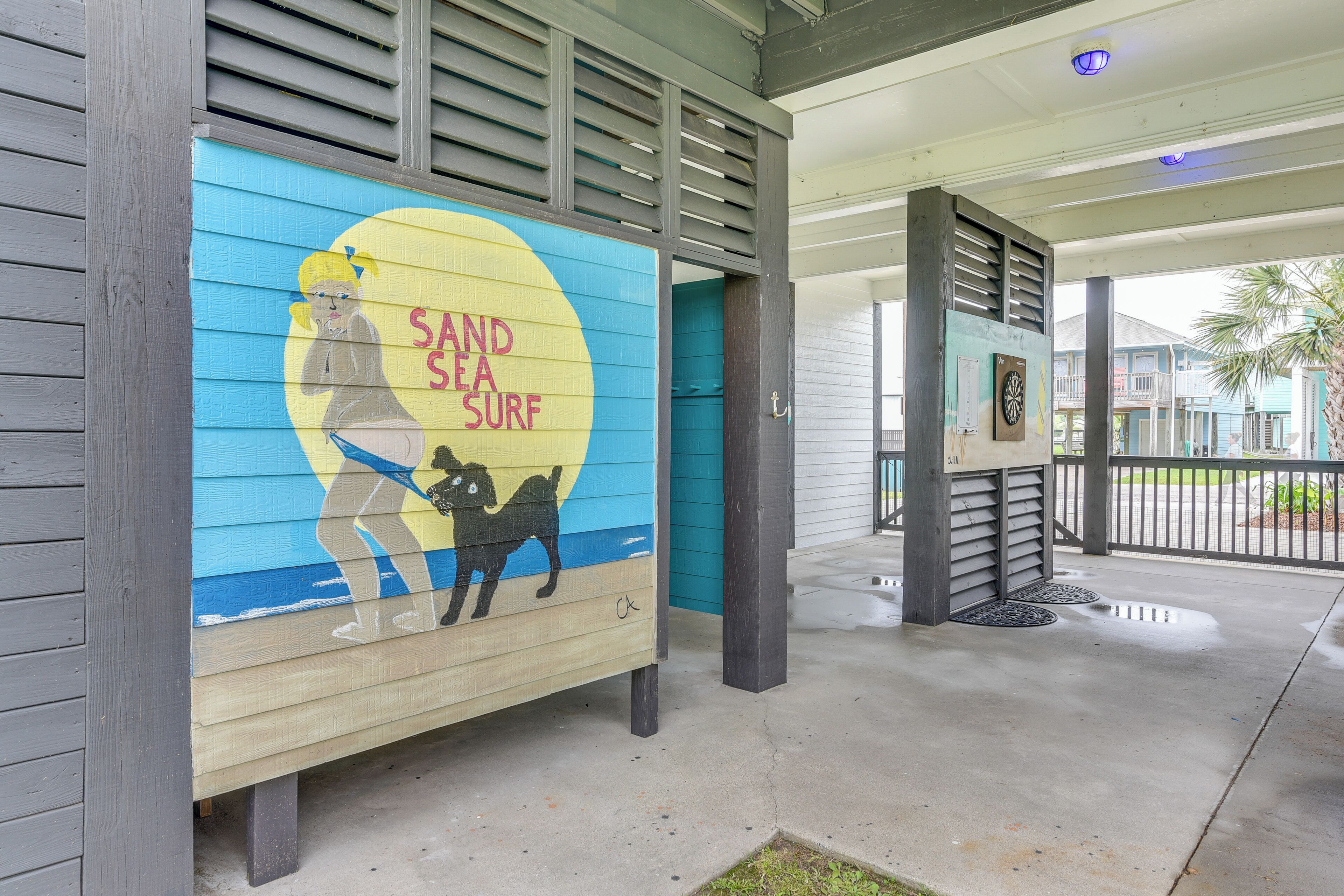 Een gevel met een muurschildering van een vrouw en een hond op een strand, een overdekte veranda en een deur met een bordje.