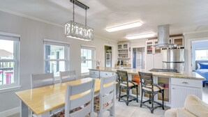 A modern kitchen with white cabinetry, a central island, and a dining area with a wooden table and chairs. - Retreat w/ Hot Tub & Fenced Yard: Walk to Beach (Port Bolivar)
