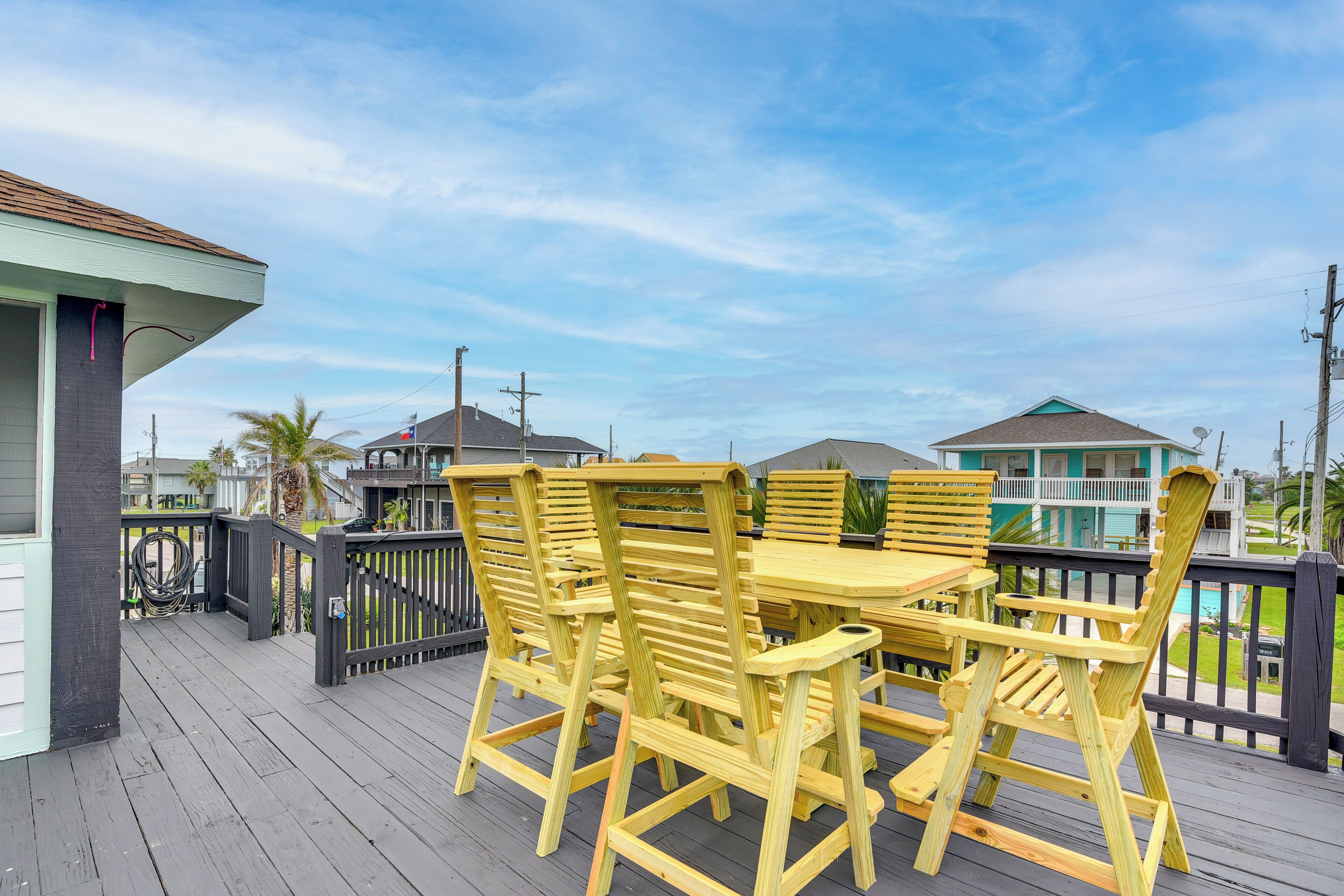 Um deck de madeira com mesa e cadeiras, com vista para uma área residencial com casas e céu claro.