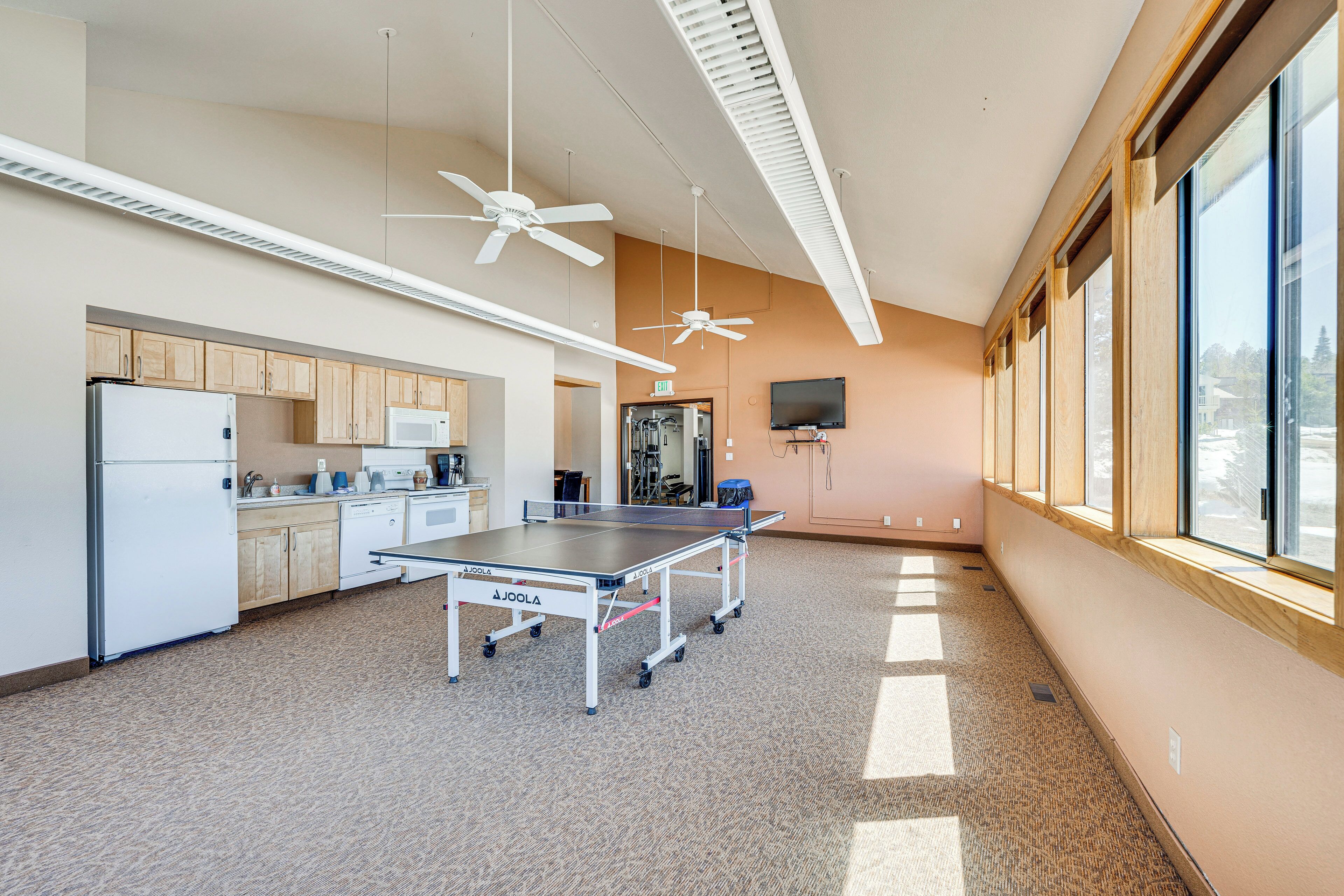 A spacious kitchen with modern appliances, a ping pong table, ceiling fans, and large windows providing natural light.