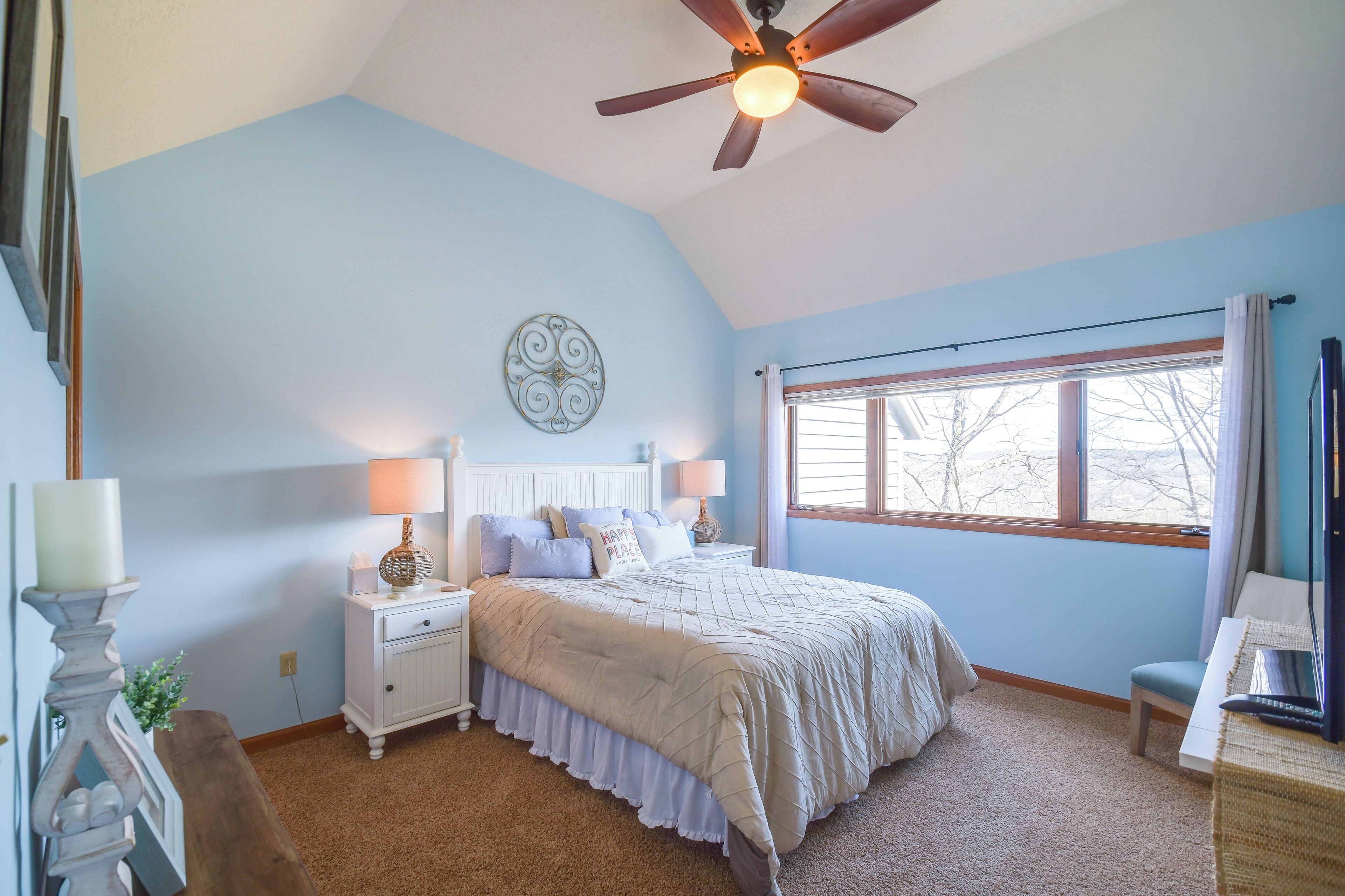A bedroom with a bed, nightstands, a ceiling fan, a window with curtains, and a wall decoration.