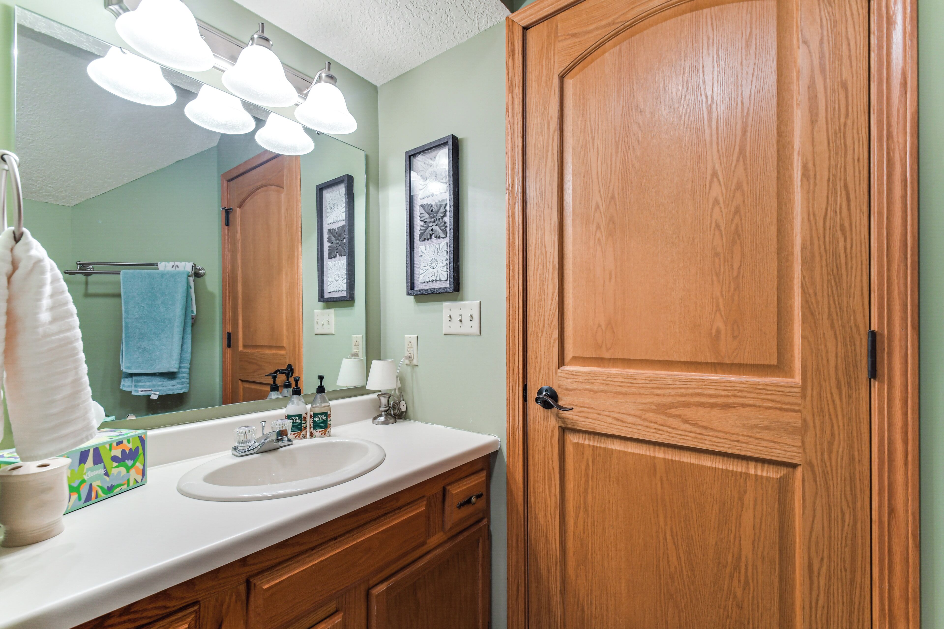 Une salle de bain avec une porte en bois, un lavabo blanc et un miroir au-dessus. Trois tableaux encadrés ornent le mur, et un porte-serviettes est fixé au mur, sur lequel est posée une serviette bleue.