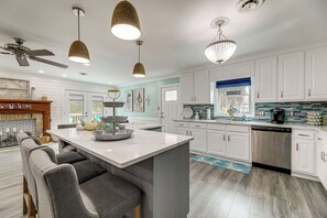 A modern kitchen with white cabinets, a central island with bar stools, pendant lighting, and a dishwasher. - Emerald Isle Beach House: Walk to the Sand! (Emerald Isle)