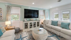 Un salon doté d’un grand téléviseur à écran plat, d’un canapé beige, d’un pouf rond en osier et d’un tapis à motifs.