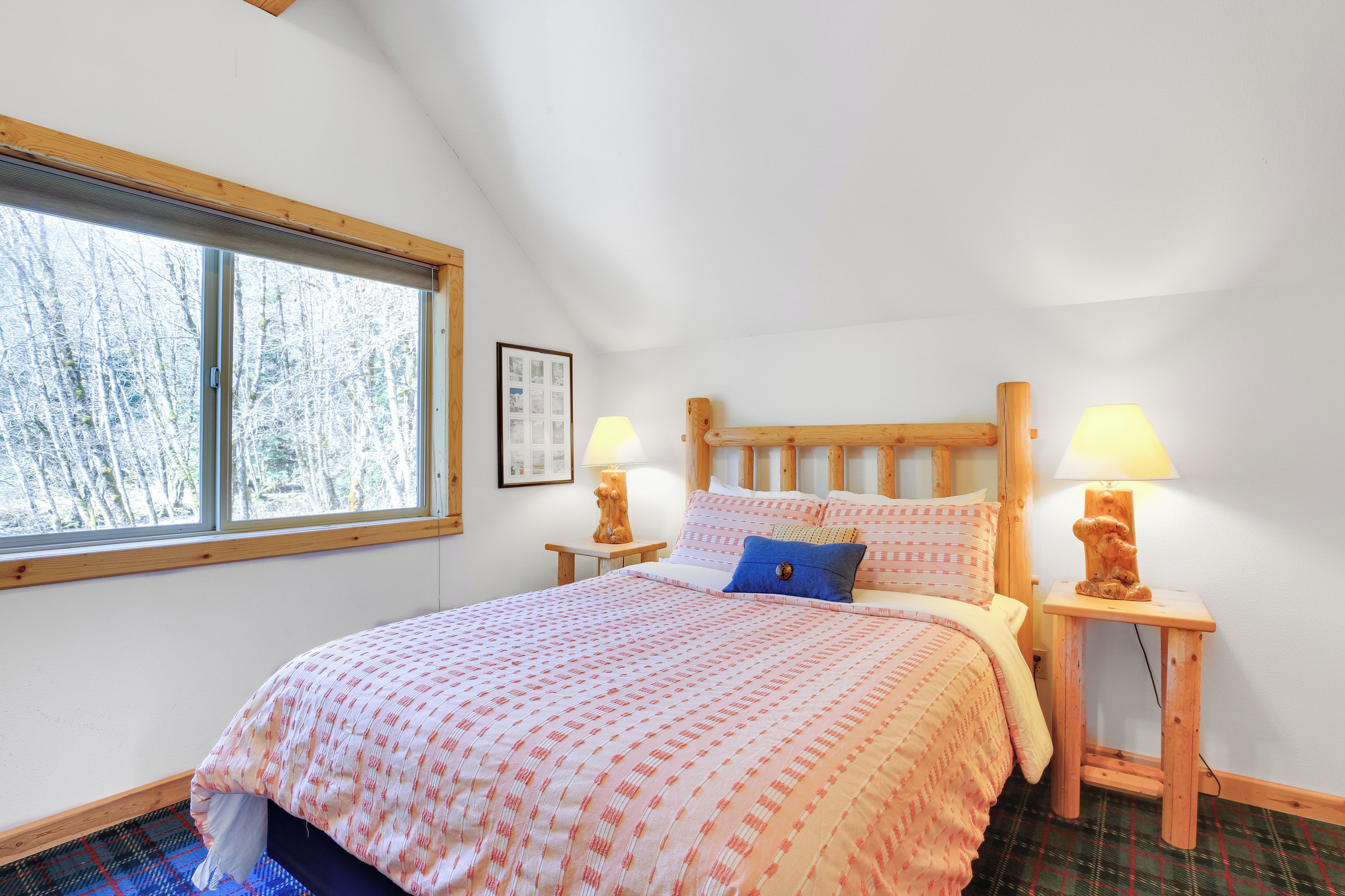 A bedroom with a large bed, two bedside tables with lamps, a window with a view of trees, and a framed picture on the wall.