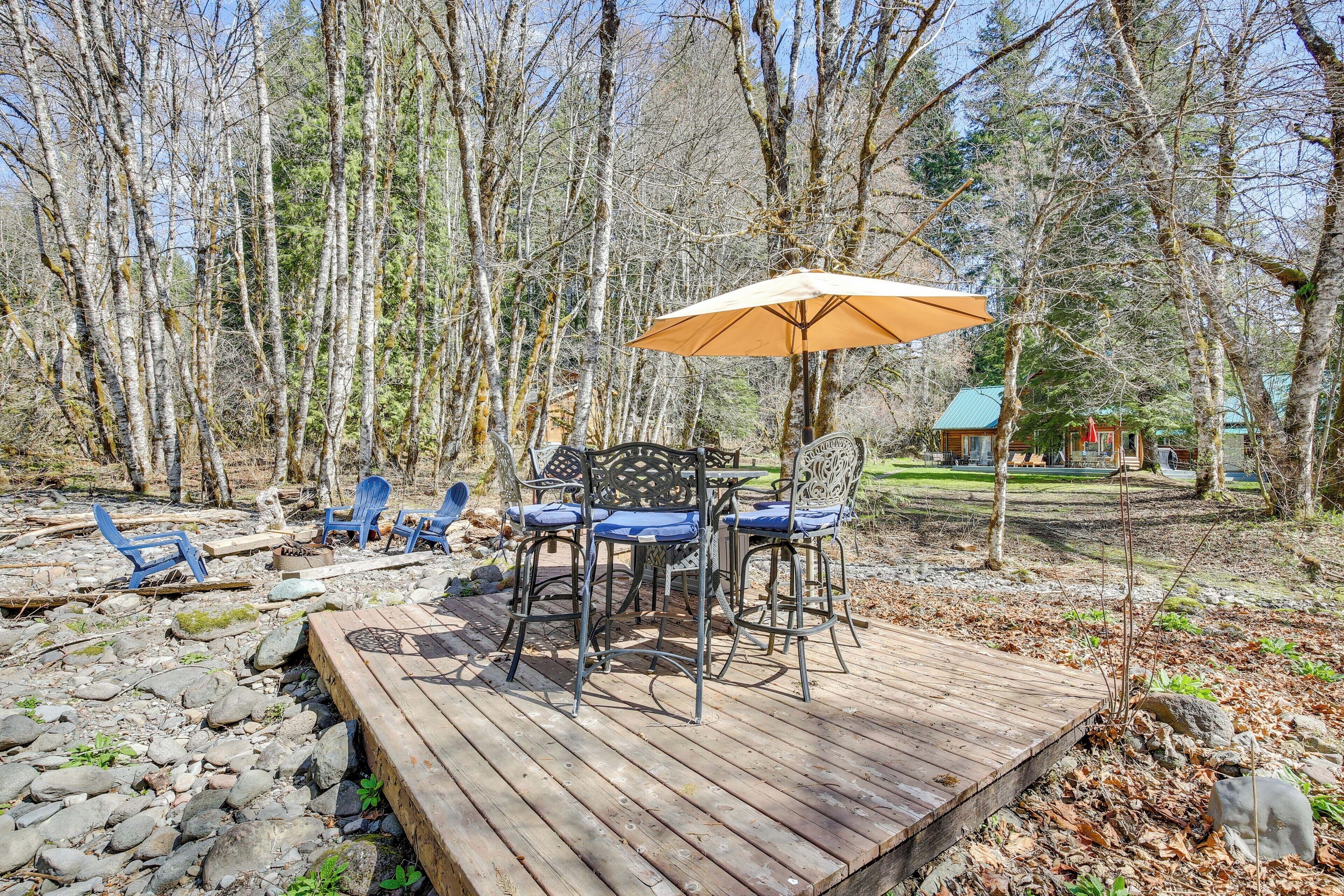 Una terraza de madera con mobiliario exterior, incluyendo una mesa y sillas, rodeada de un área boscosa.