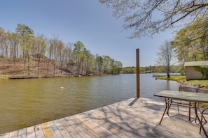 A wooden deck with a table and chairs overlooking a calm river, surrounded by trees and a small building.