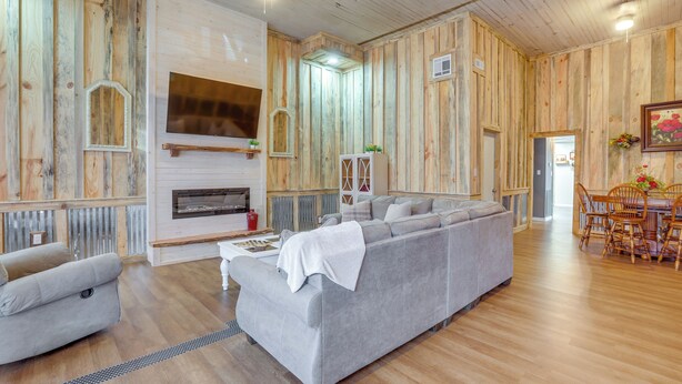 A spacious living room with a grey sectional sofa, a fireplace, wooden walls, and a dining area with wooden chairs and a table.
