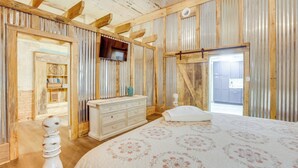 A bedroom with a wooden ceiling, a bed with a floral bedspread, a dresser, and a television on the wall.