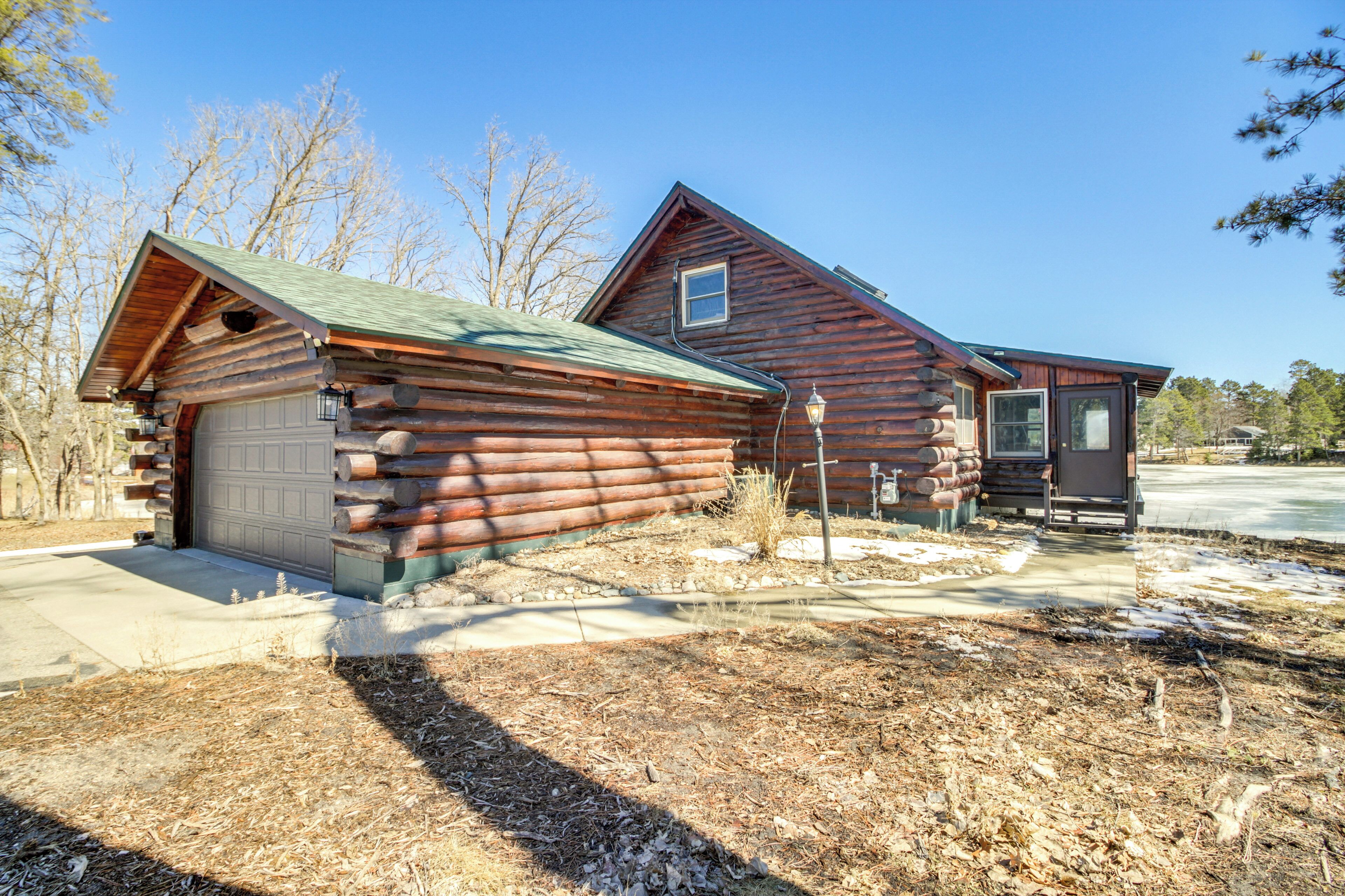 A log cabin with a garage and a small attached structure, surrounded by a yard with sparse vegetation and a clear sky.