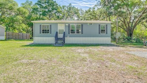 A single-story mobile home with a front porch and a small deck, surrounded by a grassy yard and trees. - 7 Mi to Crystal River: Home w/ Yard & Fire Pit! (Citrus Springs)