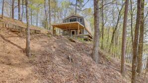 A cabin with a large deck and a covered porch, surrounded by trees and a dirt path. - Lakefront Delta Cabin Rental w/ Boat Dock + Deck! (Delta)