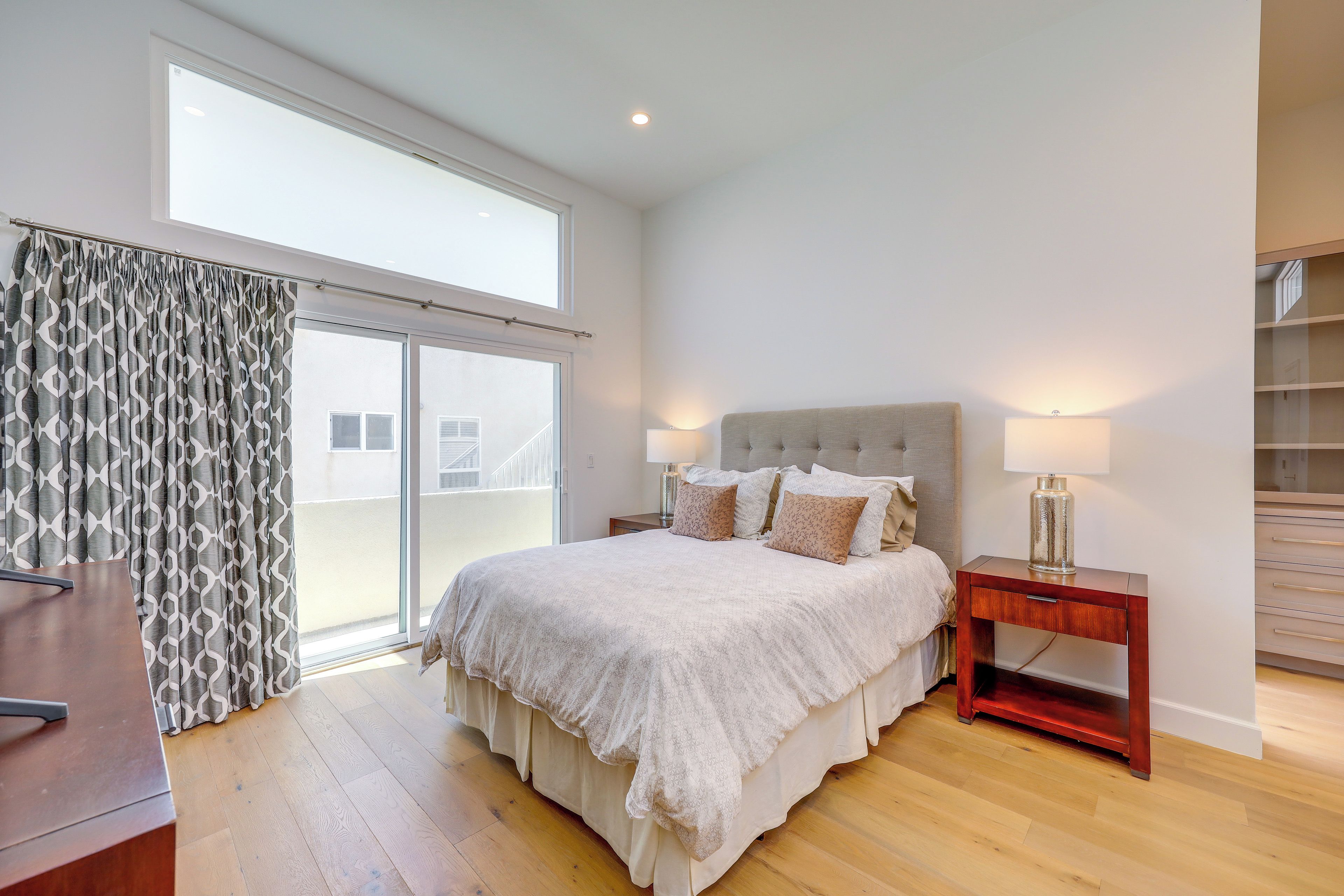 A bedroom with a large bed, a nightstand, a desk, and a view through a sliding door.