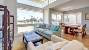 A spacious living area with a large window, a sofa, a dining table, and a coffee table. - Sleek Oxnard Townhouse w/ Balconies: Walk to Beach (Oxnard)