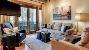 A modern living room with a large flat-screen TV, a grey sofa, a black leather armchair, and a glass coffee table.