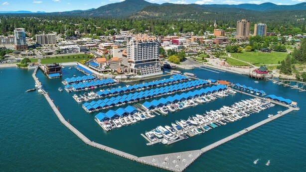 An aerial view of a marina with numerous boats docked, a large building with a prominent balcony, and a backdrop of mountains and urban development.