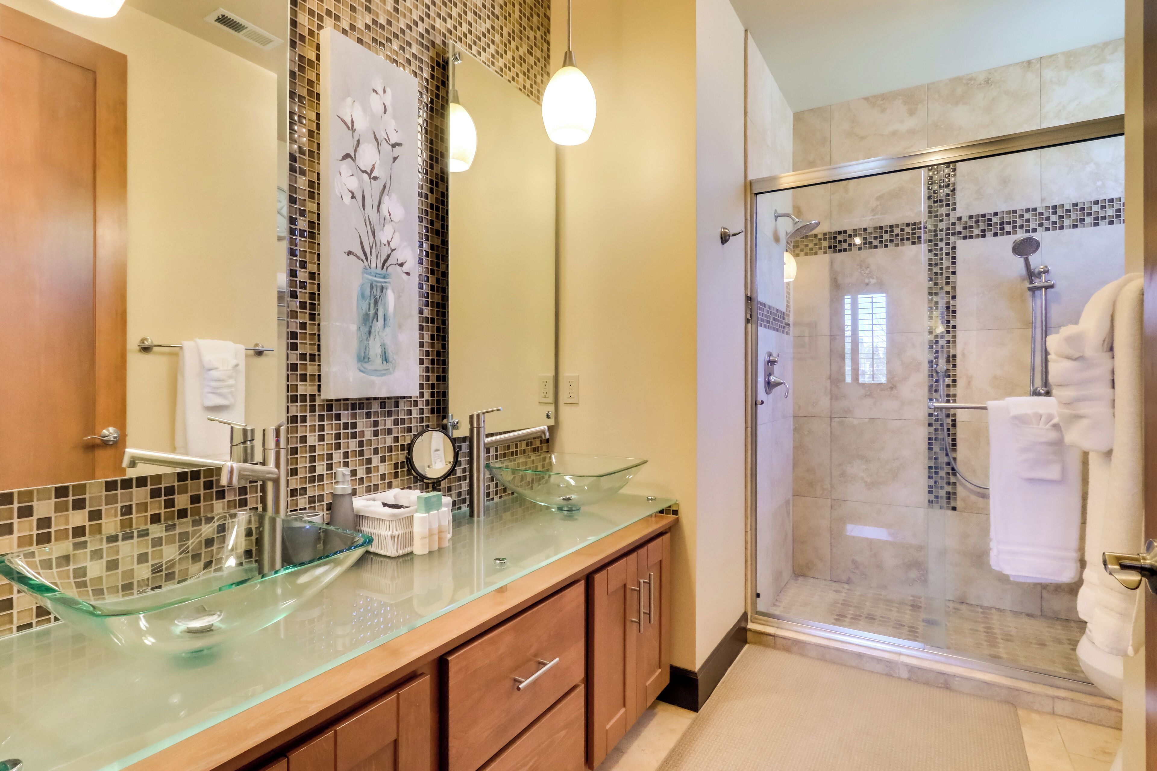 A modern bathroom with a glass vessel sink, wooden cabinets, a glass-enclosed shower, and a tiled backsplash.