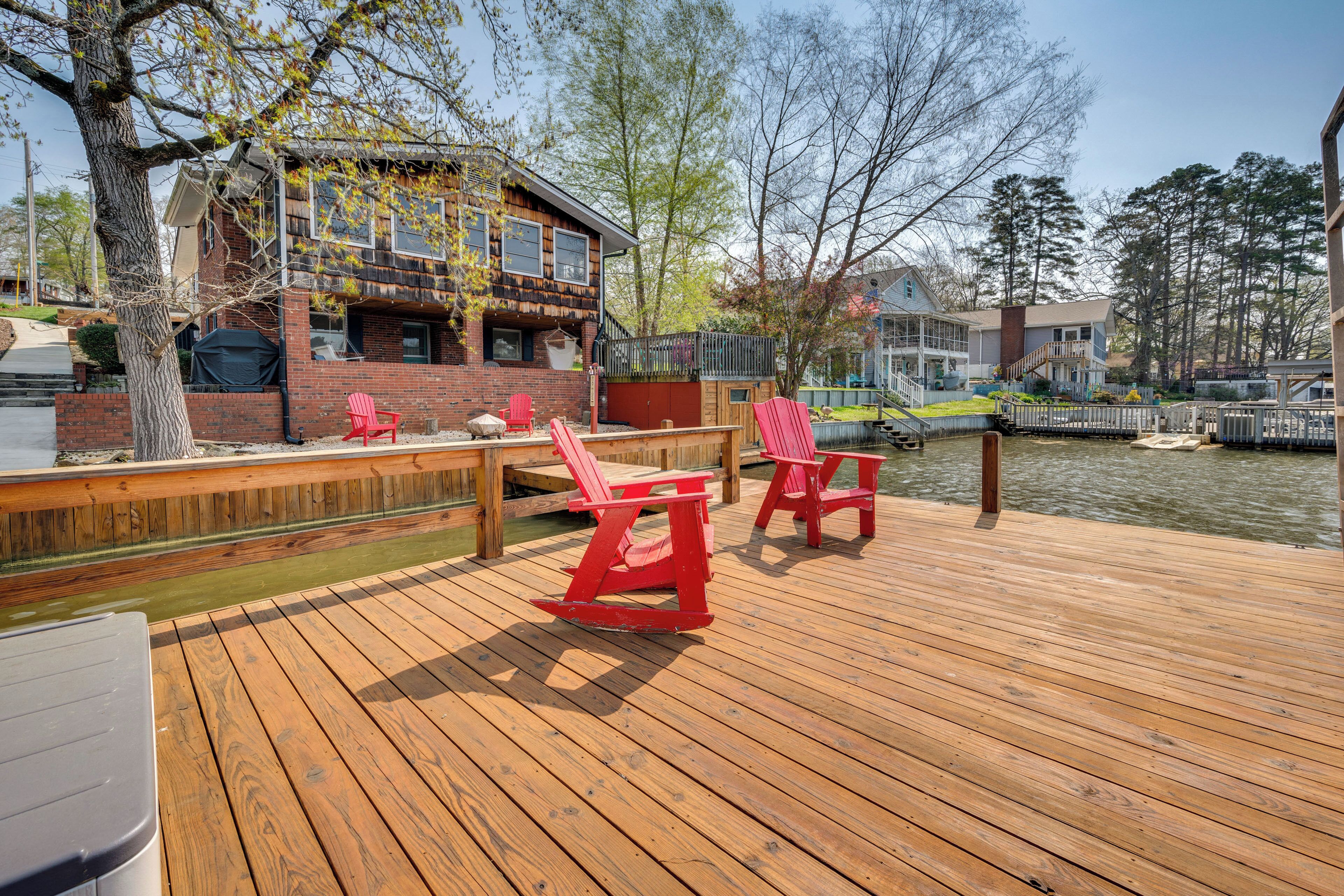 Une terrasse en bois avec des fauteuils Adirondack rouges, un bâtiment en briques et une jetée qui s’avance dans l’eau.