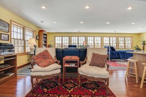 A cozy living room with two armchairs, a coffee table, and a sofa. There are large windows, a bookshelf, and a rug. - Waterfront Forest Lake Studio w/ Shared Dock! (Forest Lake)
