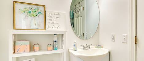Un baño con un lavabo blanco, un espejo, una obra de arte enmarcada y un estante con artículos de aseo.