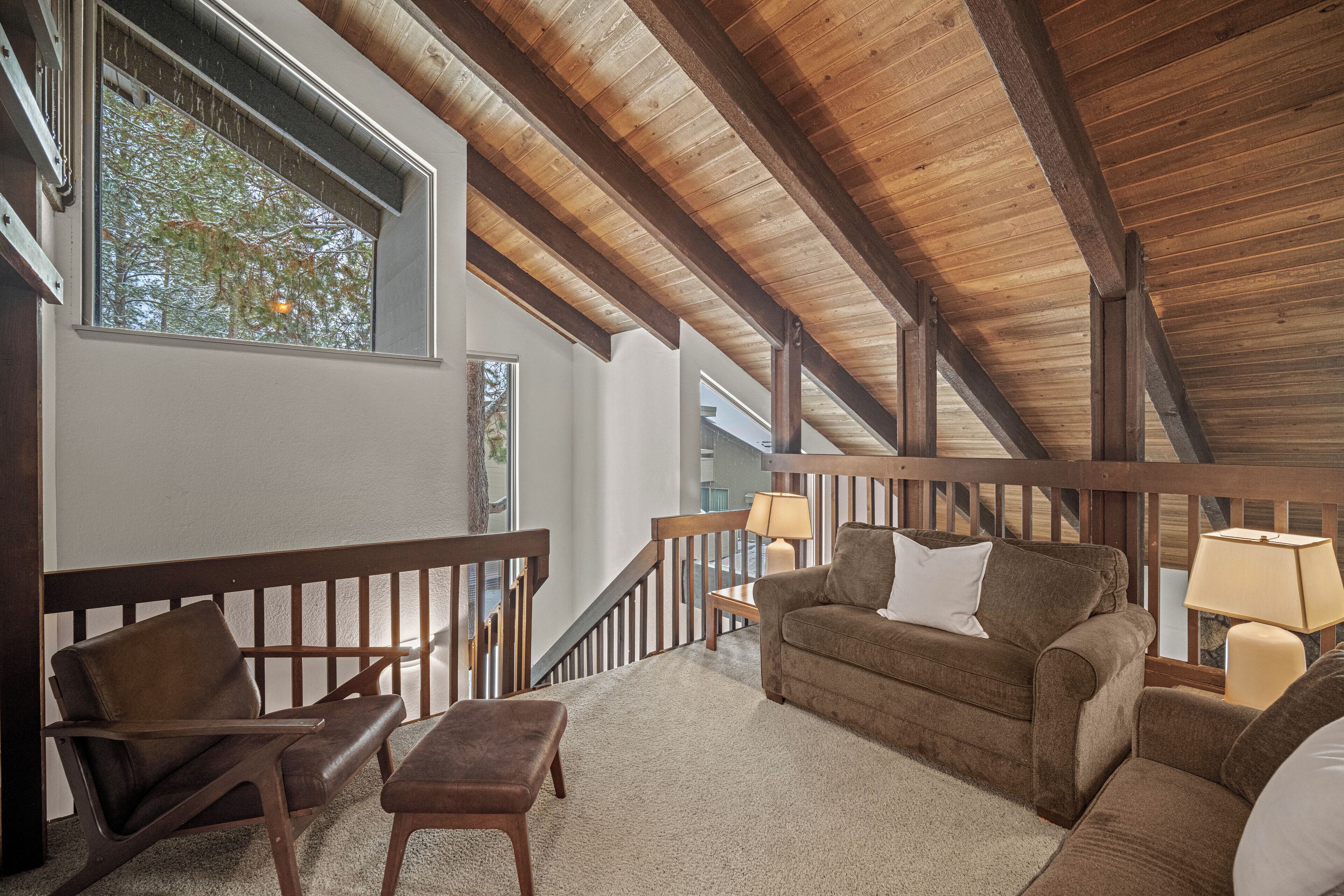 Un loft spacieux avec de hauts plafonds, des poutres apparentes, un balcon doté d’une balustrade et un coin salon confortable avec un canapé et des fauteuils.