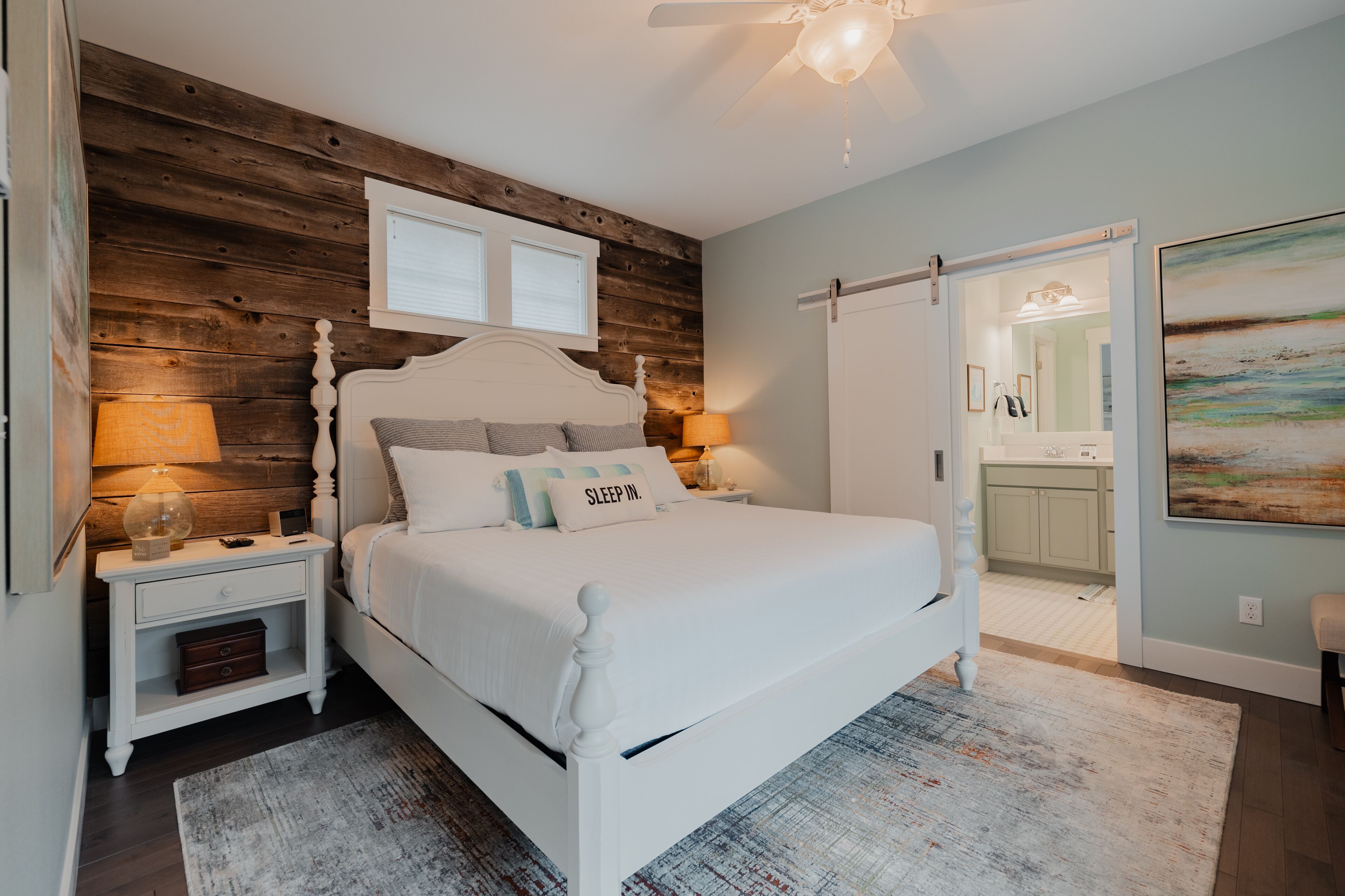 Un dormitorio con una pared decorada en madera, una cama blanca, dos lámparas de noche, una mesita de noche con cajón y un cuadro grande en la pared.