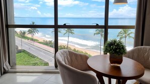 A room with a large window offering a view of the beach and ocean. There is a round table with a potted plant and two chairs. - Flat Frente Mar R608 (Maceió)