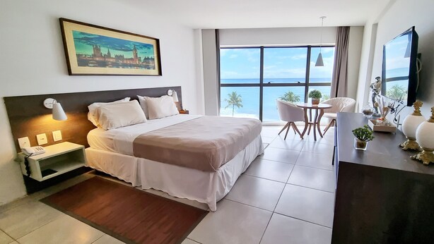 A hotel room with a large bed, a desk with a chair, a television, and a view of the ocean. - Flat Frente Mar R608 (Maceió)
