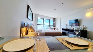 A modern hotel room with a bed, a TV, and a dining area with plates and wine glasses. - Flat Frente Mar R608 (Maceió)