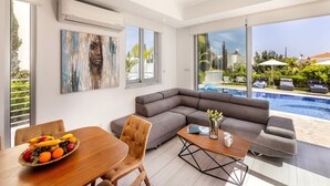 A modern living room with a pool view, a dining area with fruit, and a gray sofa. - 3 bedroom villa With Pool (Protaras)