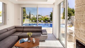 A modern living room with a sectional sofa, a coffee table with a magazine and vase, and a view of a swimming pool and outdoor seating area. - 3 bedroom villa With Pool (Protaras)