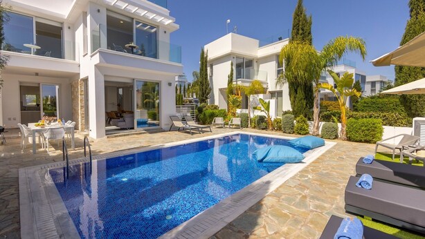 A modern poolside area with a clear blue pool, lounge chairs, and a two-story building with large windows. - 3 bedroom villa With Pool (Protaras)