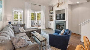 A spacious living room with a grey sofa, a blue armchair, and a coffee table. There is a fireplace, a television, and a ceiling fan. - Purple Pearl: Shared Pool with Lazy River (Kill Devil Hills)