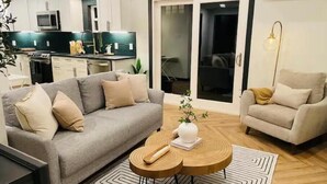 Un salon moderne avec un canapé gris, une table basse ronde en bois et un fauteuil beige. On aperçoit, au fond, une cuisine avec des meubles blancs et un four à micro-ondes.