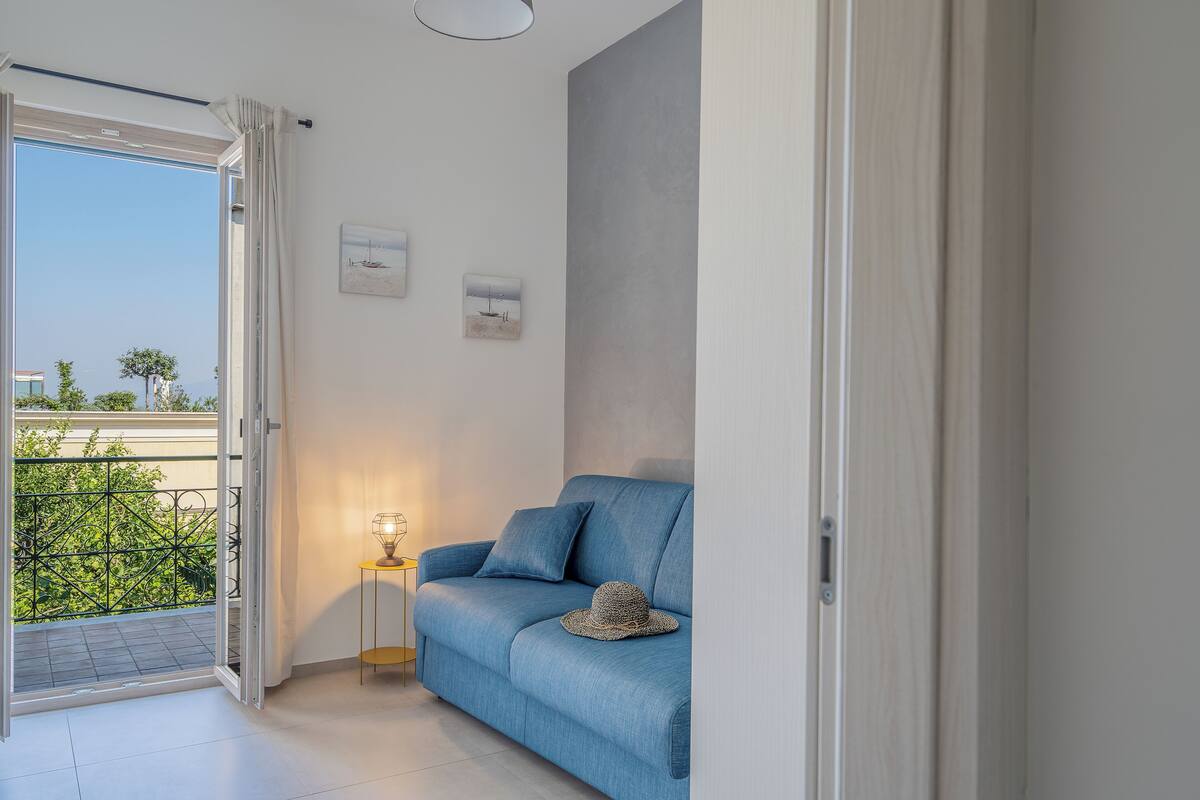 A modern living room with a blue sofa, a small table with a lamp, and a balcony with a view of greenery and buildings.