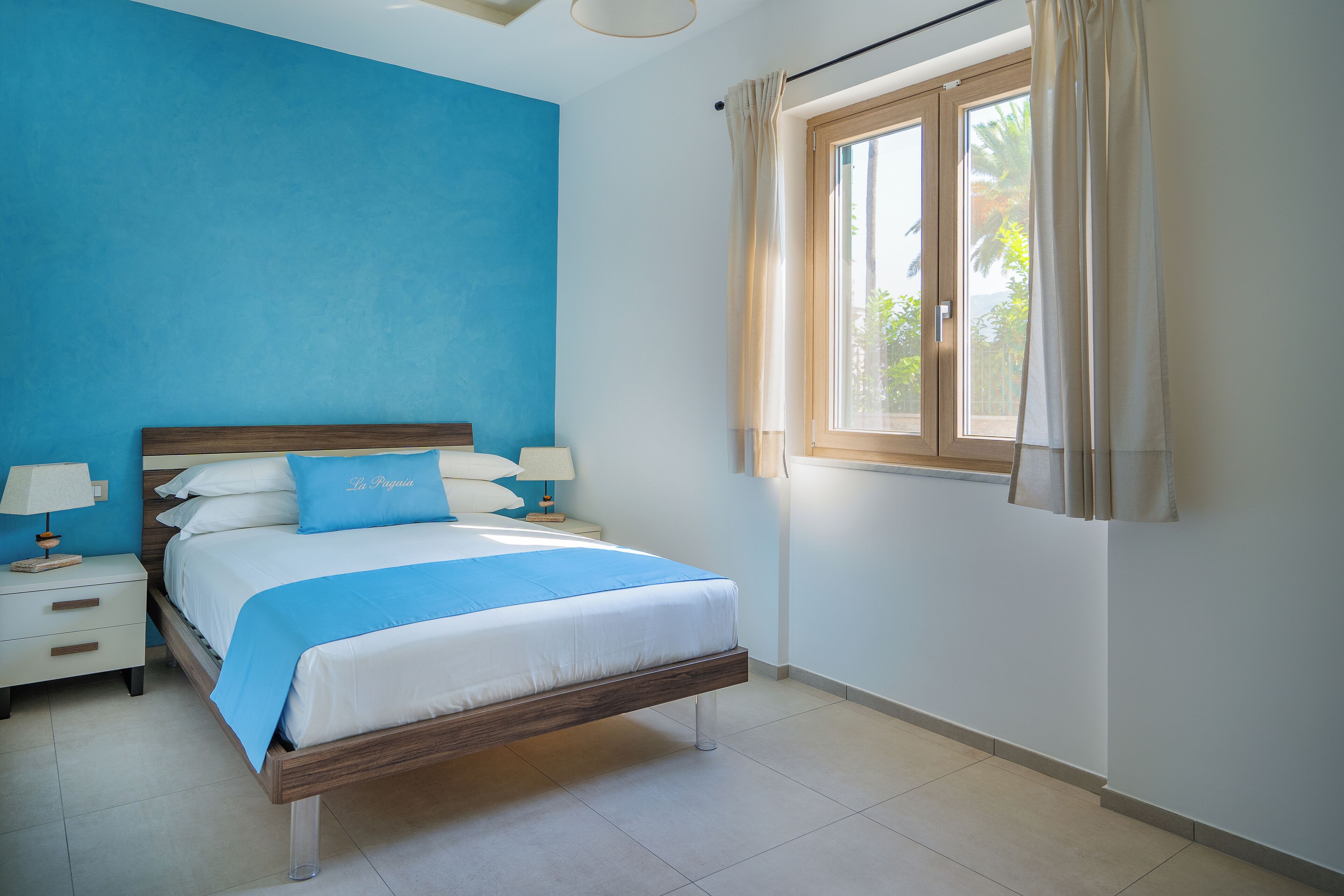 Un dormitorio con cama, mesitas de noche y ventana con cortinas.