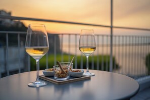 To glas hvidvin på et bord med skåle med nødder og en ske, med udsigt til en balkon med gelænder og en solnedgang.