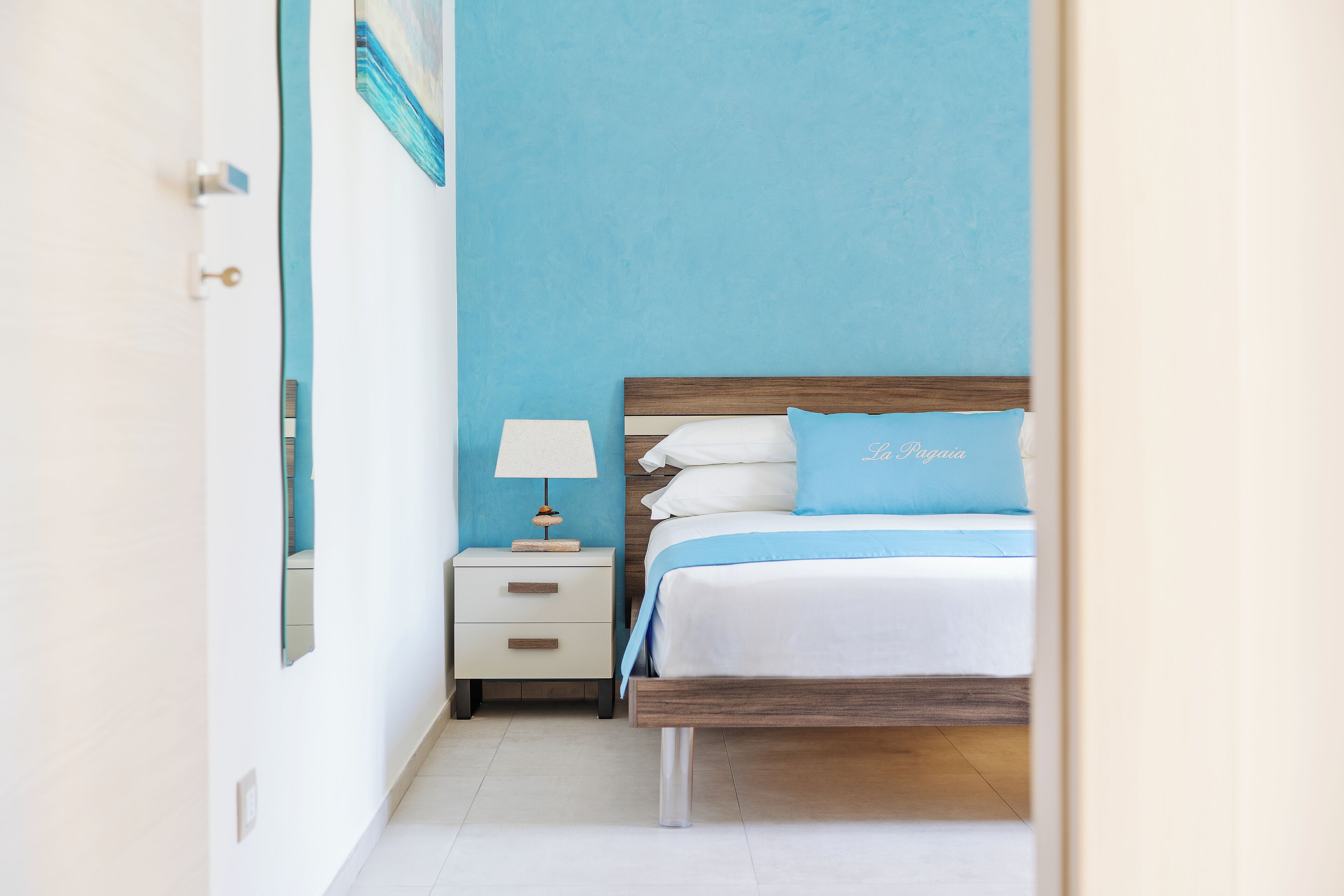 A bedroom with a bed, a nightstand, a lamp, and a wall with a painting.