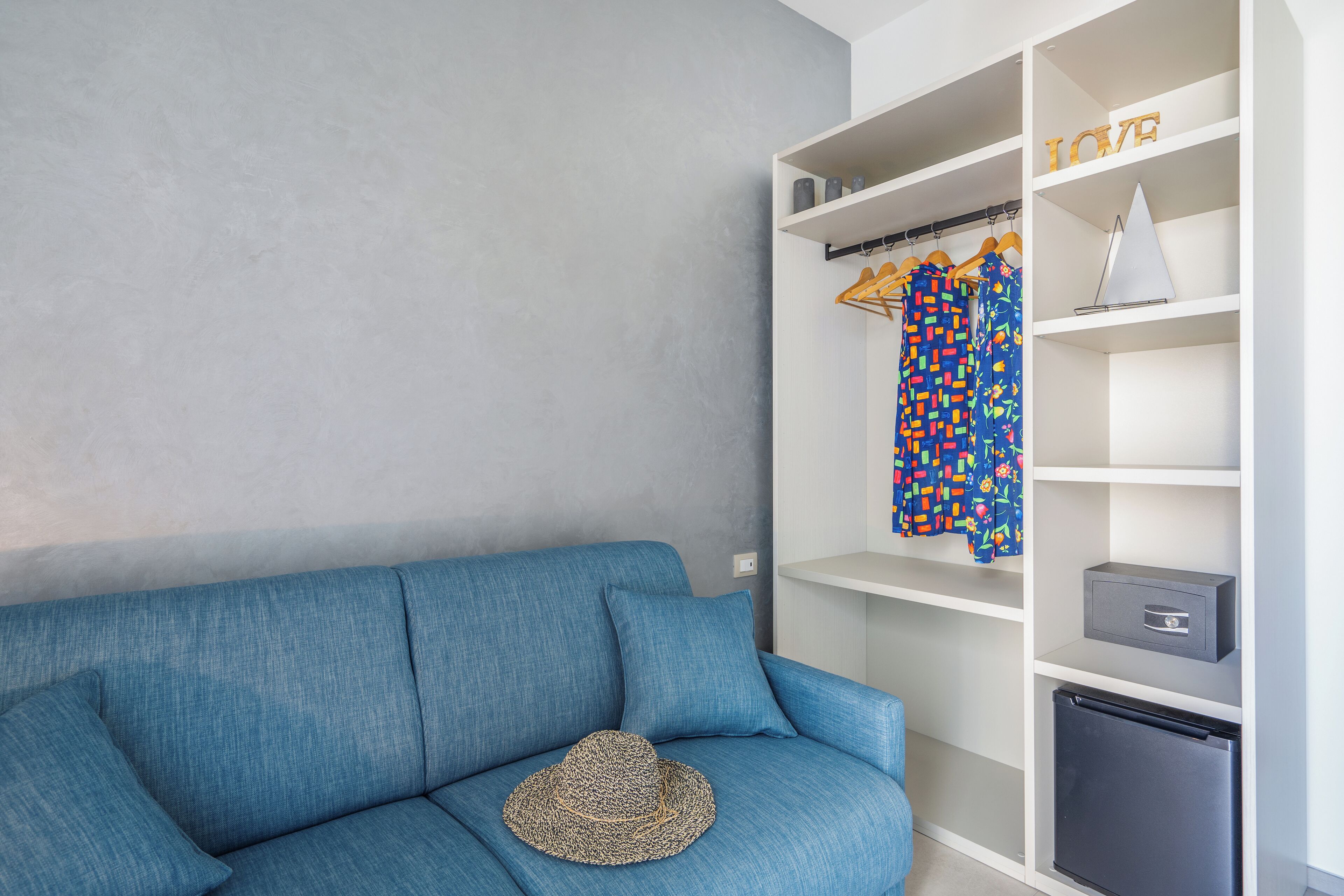Una habitación con un sofá azul, un sombrero en el asiento y un armario blanco con estantes y un refrigerador pequeño.