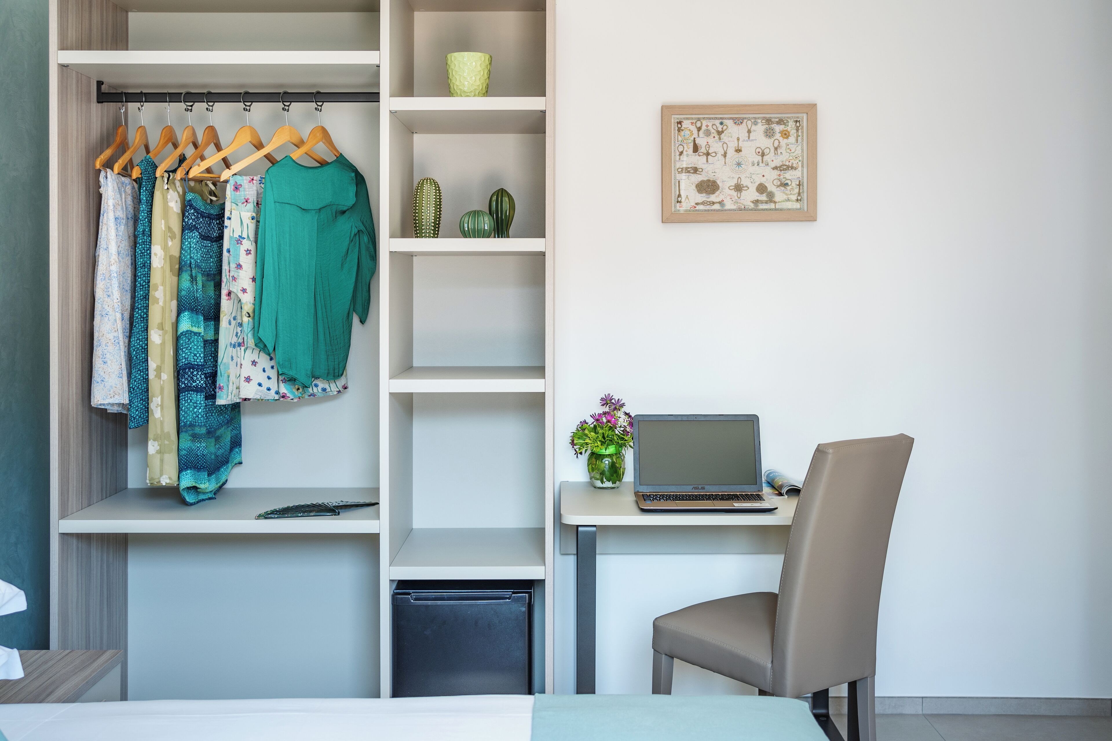 A neatly organized room with a bed, a desk with a laptop, a chair, a wardrobe with clothes, a small cactus collection, and a framed picture on the wall.
