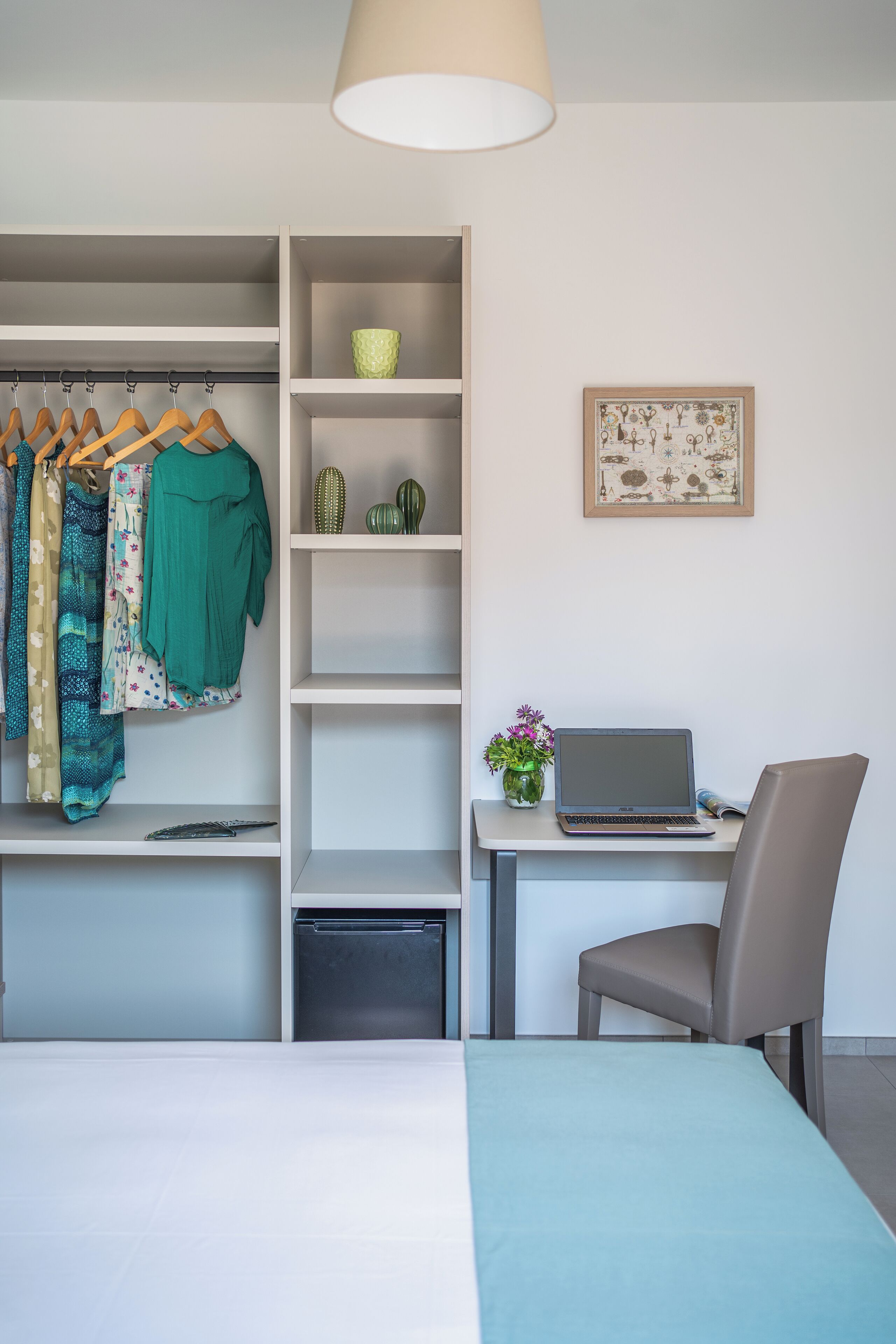 A bedroom with a bed, a desk with a laptop, a chair, a wardrobe with clothes, a shelf with decorative items, and a vase with flowers.