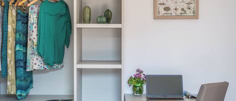 A bedroom with a bed, a desk with a laptop, a chair, a wardrobe with clothes, a shelf with decorative items, and a vase with flowers.