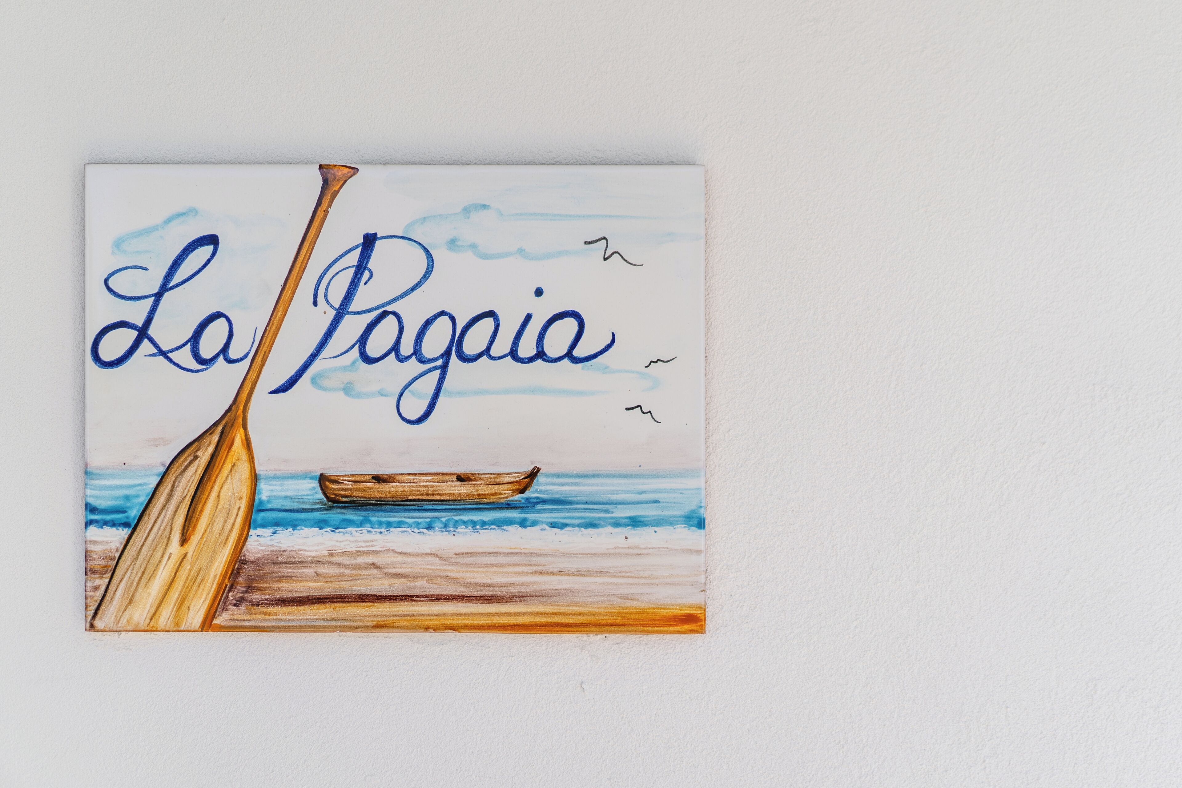Ein Gemälde von einem hölzernen Boot am Strand, mit dem Schriftzug 'La Pagaaia' in Blau.