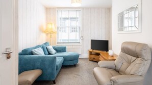 Un salon avec un canapé bleu, un fauteuil beige, un téléviseur posé sur un meuble en bois et une fenêtre avec des stores.