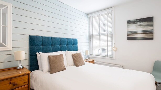 Une chambre à coucher avec une tête de lit bleue recouverte de tissu, du linge de lit blanc, une table de chevet en bois avec une lampe et une fenêtre avec des stores.
