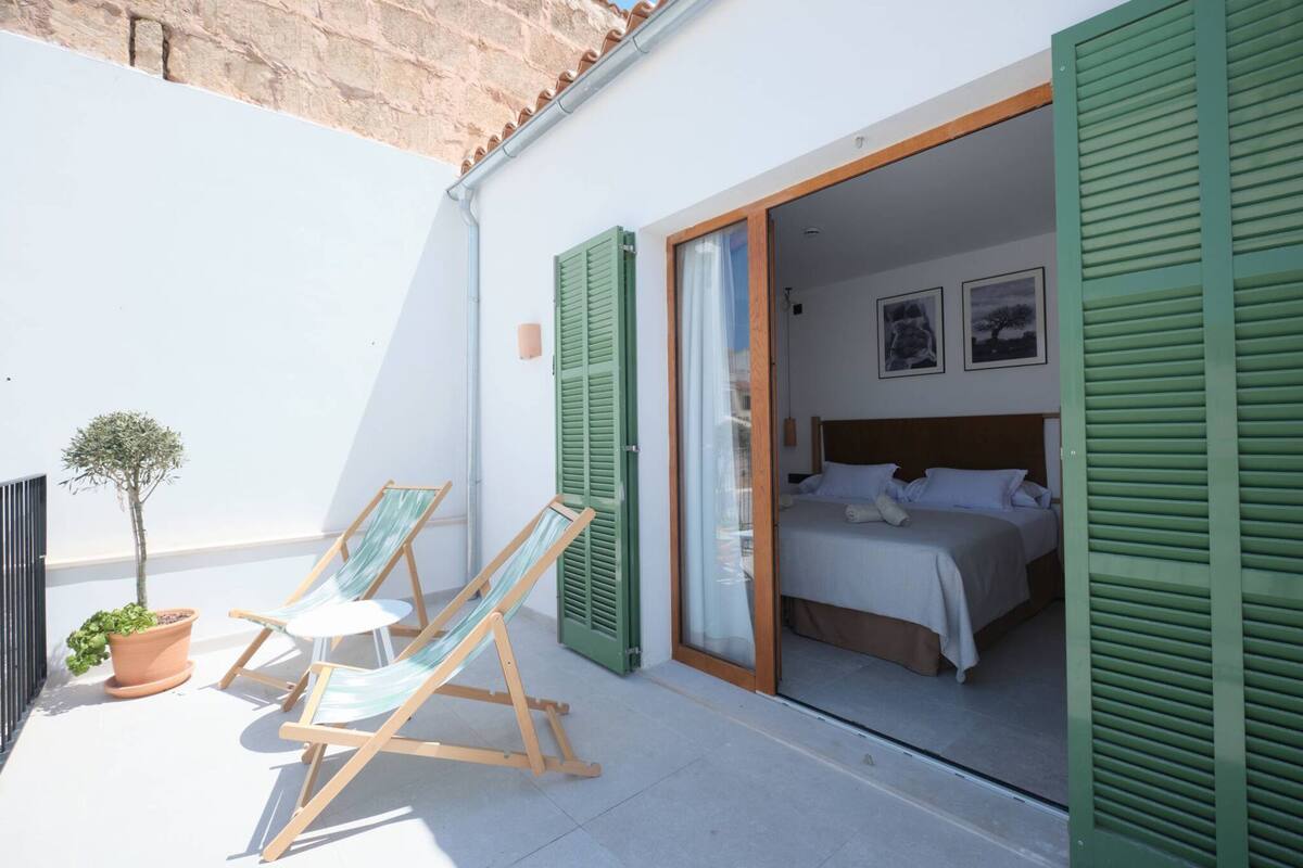A bedroom with a bed, a small balcony with a potted plant, and two lounge chairs.