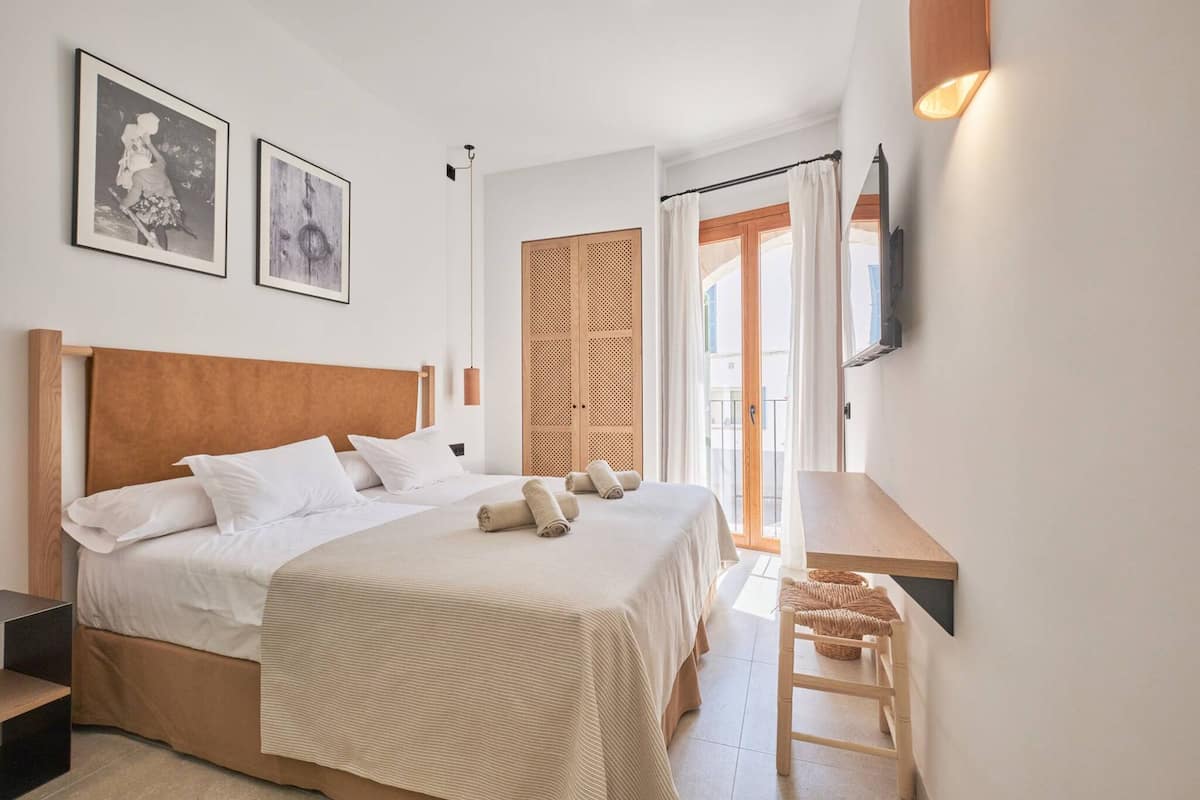 A bedroom with a large bed, a wooden headboard, a bedside table, a TV mounted on the wall, and a door leading to a balcony.