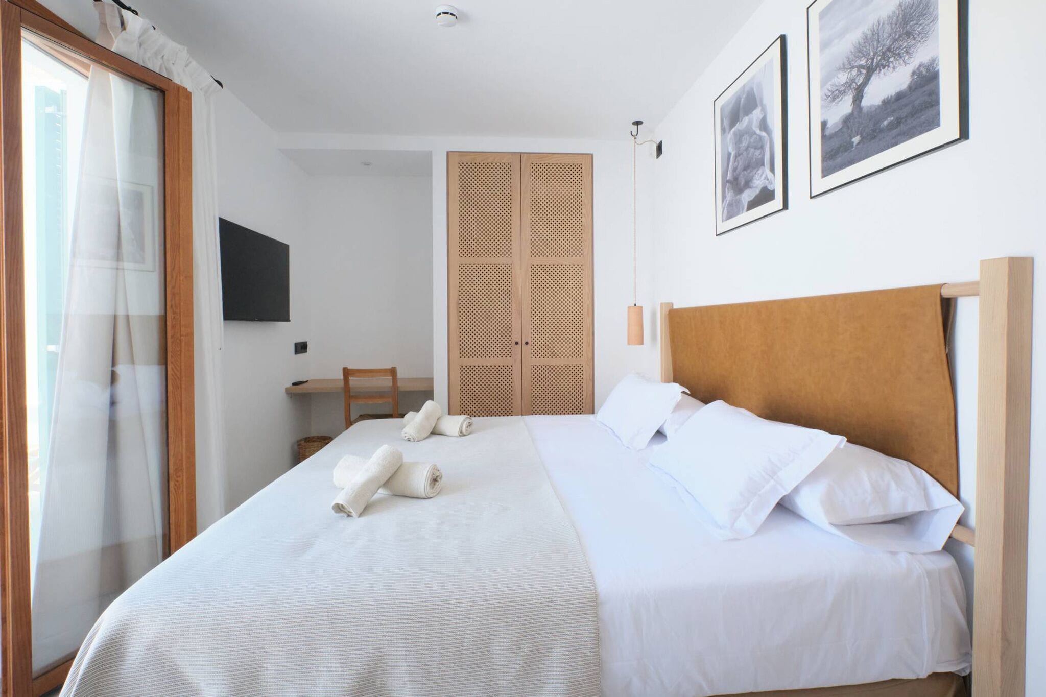 Una habitación de hotel con una cama grande, cabecera de madera, armario de madera, una silla y un ventanal con cortinas.