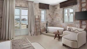 A bathroom with a stone wall, a bathtub, a ceiling fan, and a view of the sea. - Horizon Suites Milos (Milos)