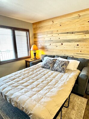 A bedroom with a bed, a grey sofa, a wooden headboard, a window with blinds, a lamp, and a rug.
